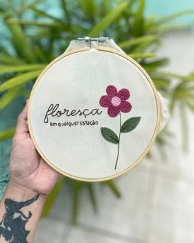 Bordado de aluna em bastidor com flor rosa, folhas verdes e a frase floresça em qualquer estação