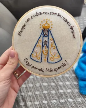 Bordado de aluna em bastidor com Nossa Senhora de manto azul, coroa dourada e uma oração