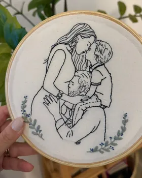 Bordado de aluna em bastidor com uma família abraçada, contornos pretos e ramos de folhas