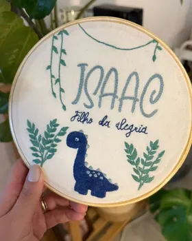 Bordado de aluna em bastidor com dinossauro azul, folhagens e a inscrição Isaac, filho da alegria